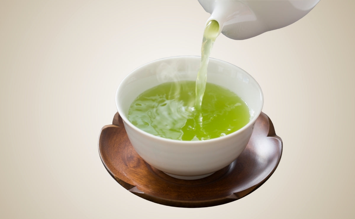Green tea being poured into a white bowl on a wooden coaster on a beige background