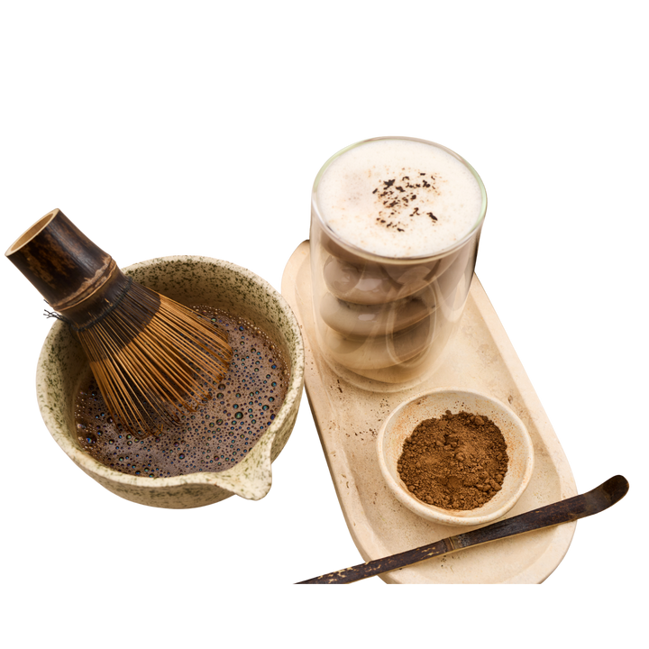 A hojicha preparation set featuring a whisk in a ceramic bowl filled with whisked hojicha powder, a glass of layered hojicha latte with foam, a small bowl of loose hojicha powder and a serving spoon