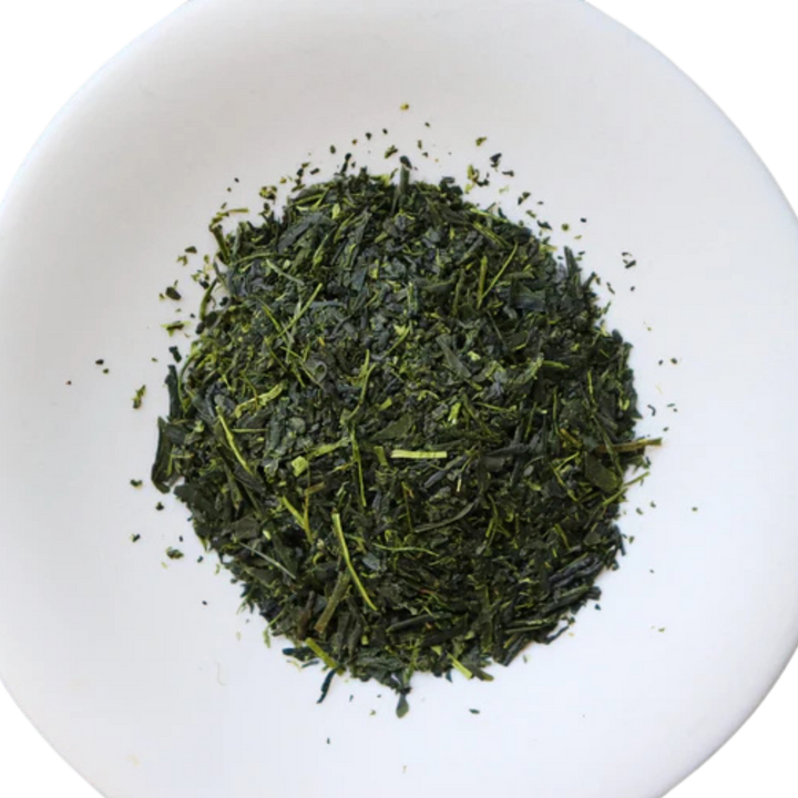 A pile of loose-leaf dark green Sencha tea leaves with a few lighter green stems, presented in a round white dish