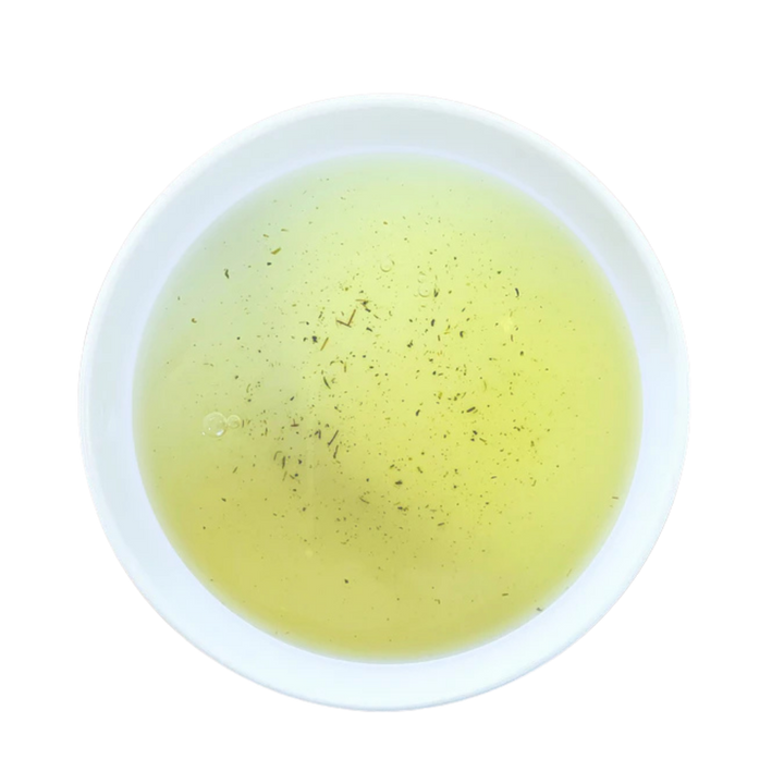 A top-down view of a white cup filled with a pale yellow tea, with small dark herb flecks at the bottom