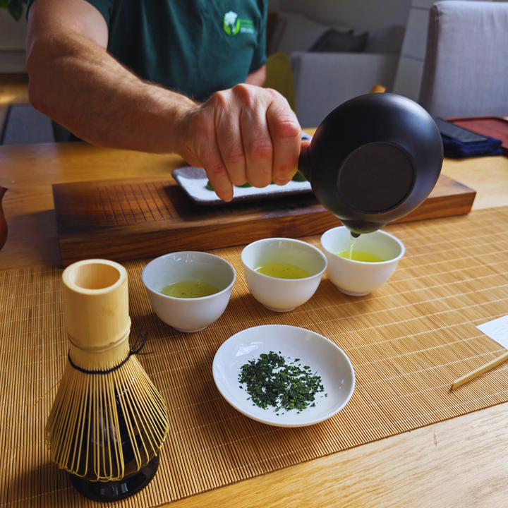 A person pouring tea from a black kettle into four small white cups on a bamboo mat, with a whisk and a small dish of loose leaf in the foreground.