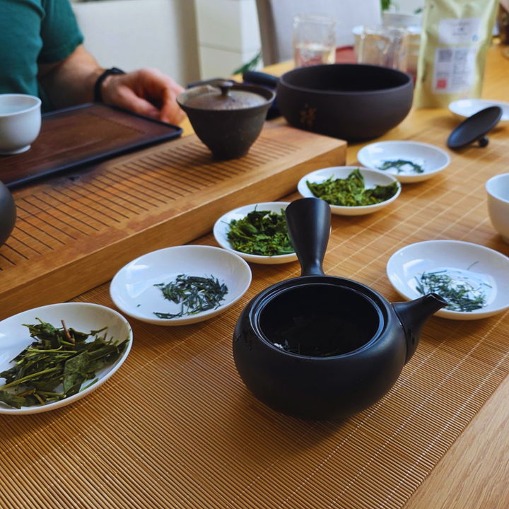 A black ceramic teapot sits on a bamboo tea table alongside small white dishes of loose leaf tea, with a person's hand visible in the background near a matching black bowl.