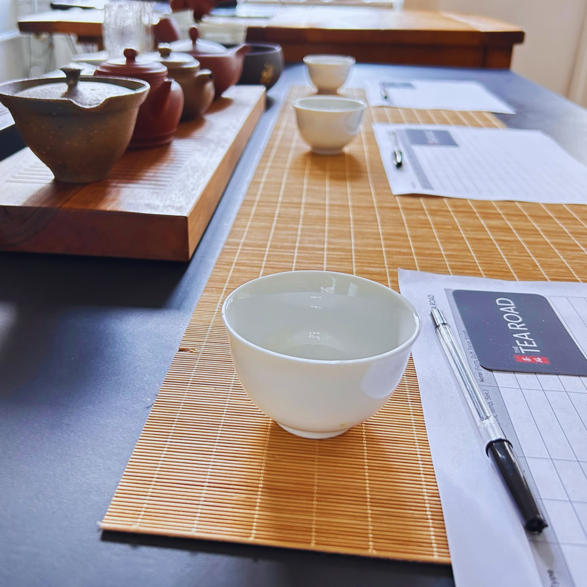 A white ceramic tea bowl sits on a bamboo placemat on a dark table, alongside a pen and a printed form with a visible TearRoad logo. More tea vessels and papers are arranged along the table in the background.