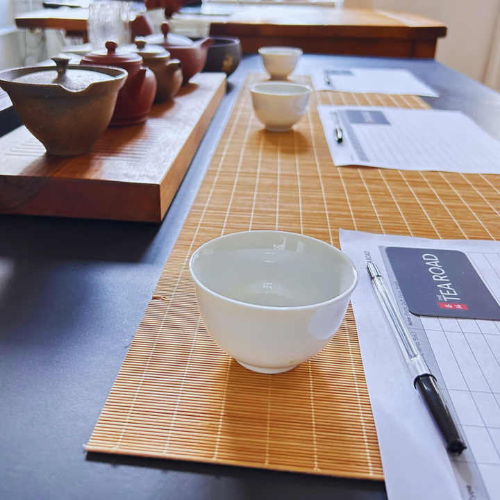 A white ceramic tea bowl sits on a bamboo placemat on a dark table, alongside a pen and a printed form with a visible TearRoad logo. More tea vessels and papers are arranged along the table in the background.