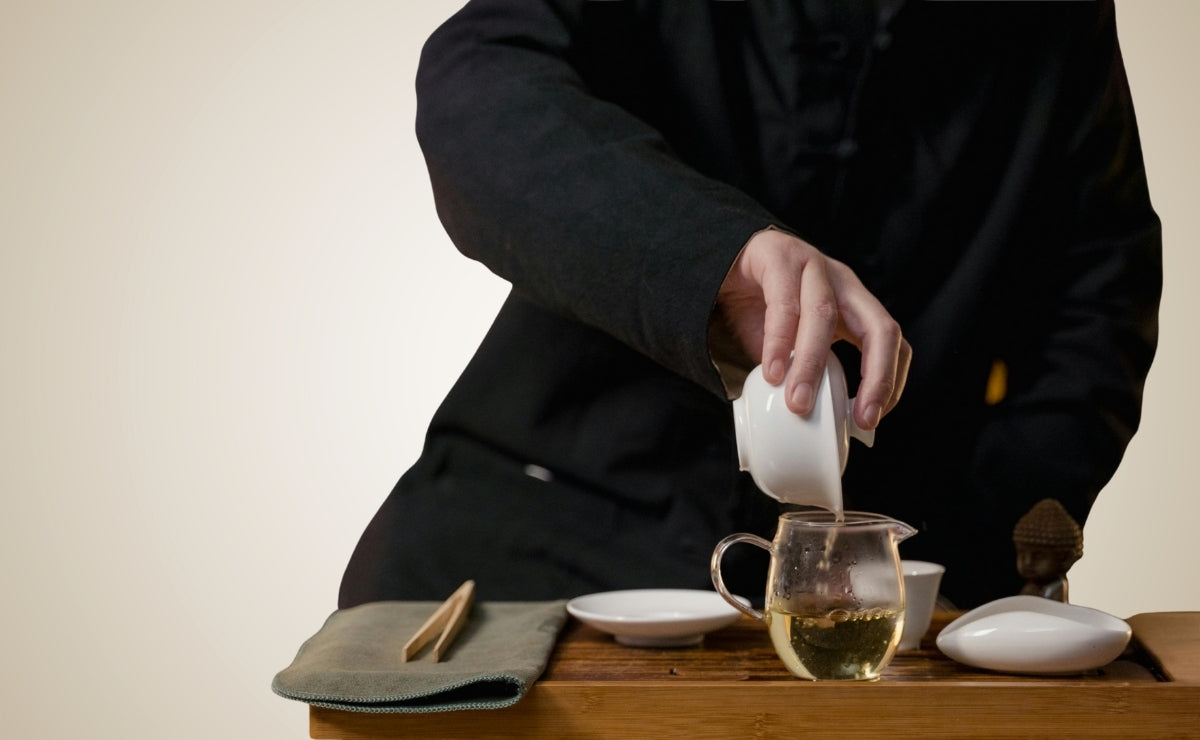 Person pouring tea from a gaiwan to a glass pitcher on a wooden tray