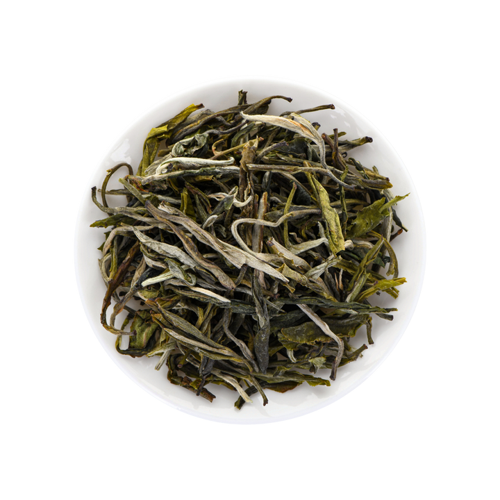 A top-down view of loose-leaf tea leaves, showing green and white buds with dark stems, presented in a small white bowl